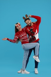 Photo of Happy young couple in Christmas sweaters, reindeer headband and funny glasses on light blue background