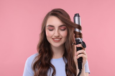 Photo of Beautiful young woman using curling hair iron on pink background