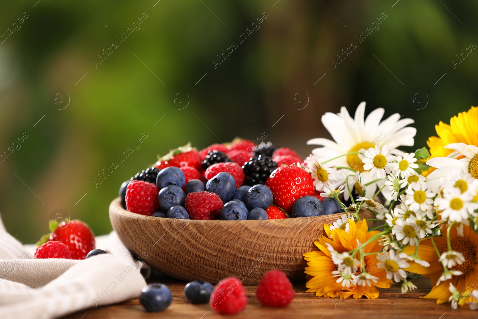 Photo of Bowl with different fresh ripe berries and beautiful flowers on table outdoors