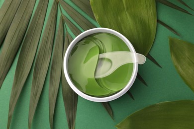 Photo of Jar of under eye patches with spoon and leaves on green background, top view. Cosmetic product