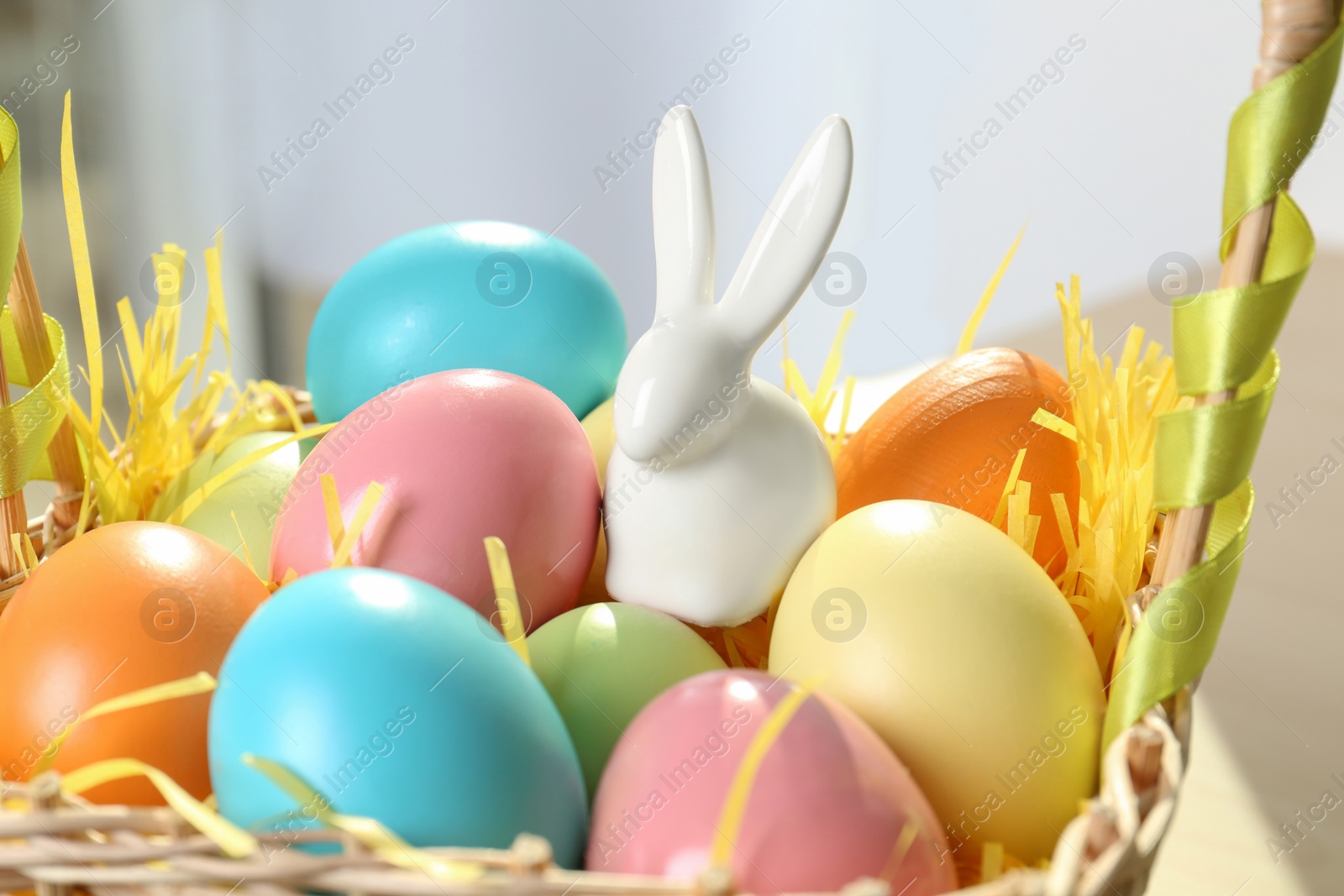 Photo of Cute ceramic Easter bunny and dyed eggs in wicker basket on table, closeup