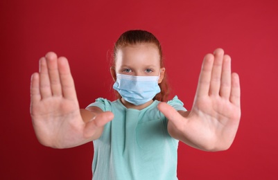 Photo of Little girl in protective mask showing stop gesture on red background. Prevent spreading of coronavirus