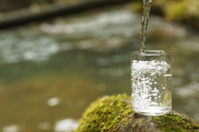 Fresh water pouring into glass on stone near river. Space for text