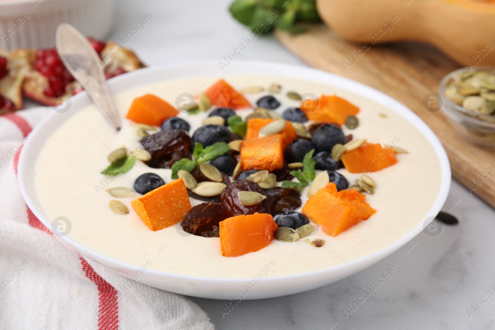 Photo of Delicious semolina pudding with blueberries, dates, pumpkin, spoon and mint in bowl on white table, closeup