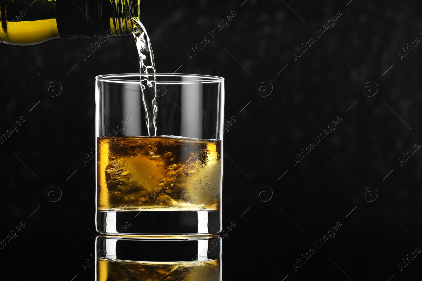 Photo of Pouring tasty whiskey from bottle into glass with ice at mirror table against black background, closeup. Space for text