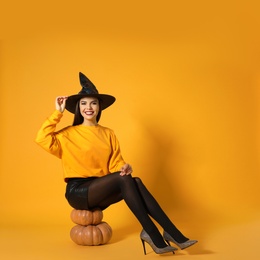 Beautiful woman wearing witch costume for Halloween party sitting on pumpkins against yellow background, space for text