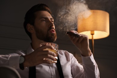Photo of Handsome man with glass of whiskey smoking cigar at home, low angle view