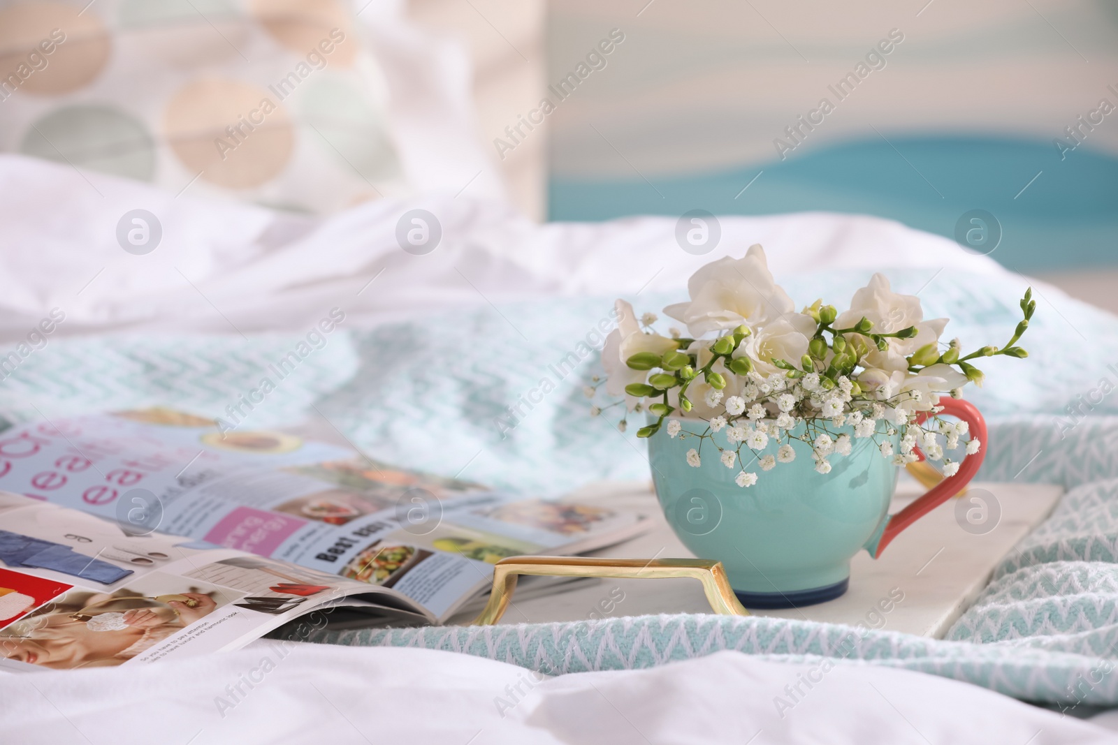 Photo of Beautiful bright flowers in cup, open magazine and tray on bed. Space for text