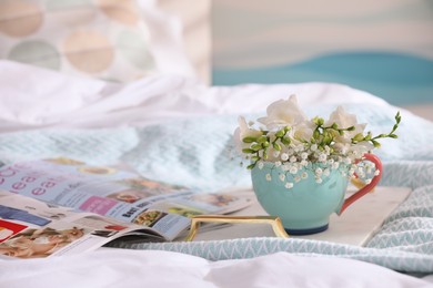 Photo of Beautiful bright flowers in cup, open magazine and tray on bed. Space for text