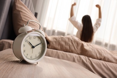 Photo of It's lazy morning o'clock. Alarm clock on bedside table and woman stretching in room, selective focus