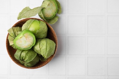 Photo of Fresh green tomatillos with husk in bowl on white tiled table, flat lay. Space for text