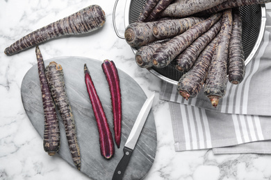 Flat lay composition with raw black carrots, knife and cutting board on white marble table