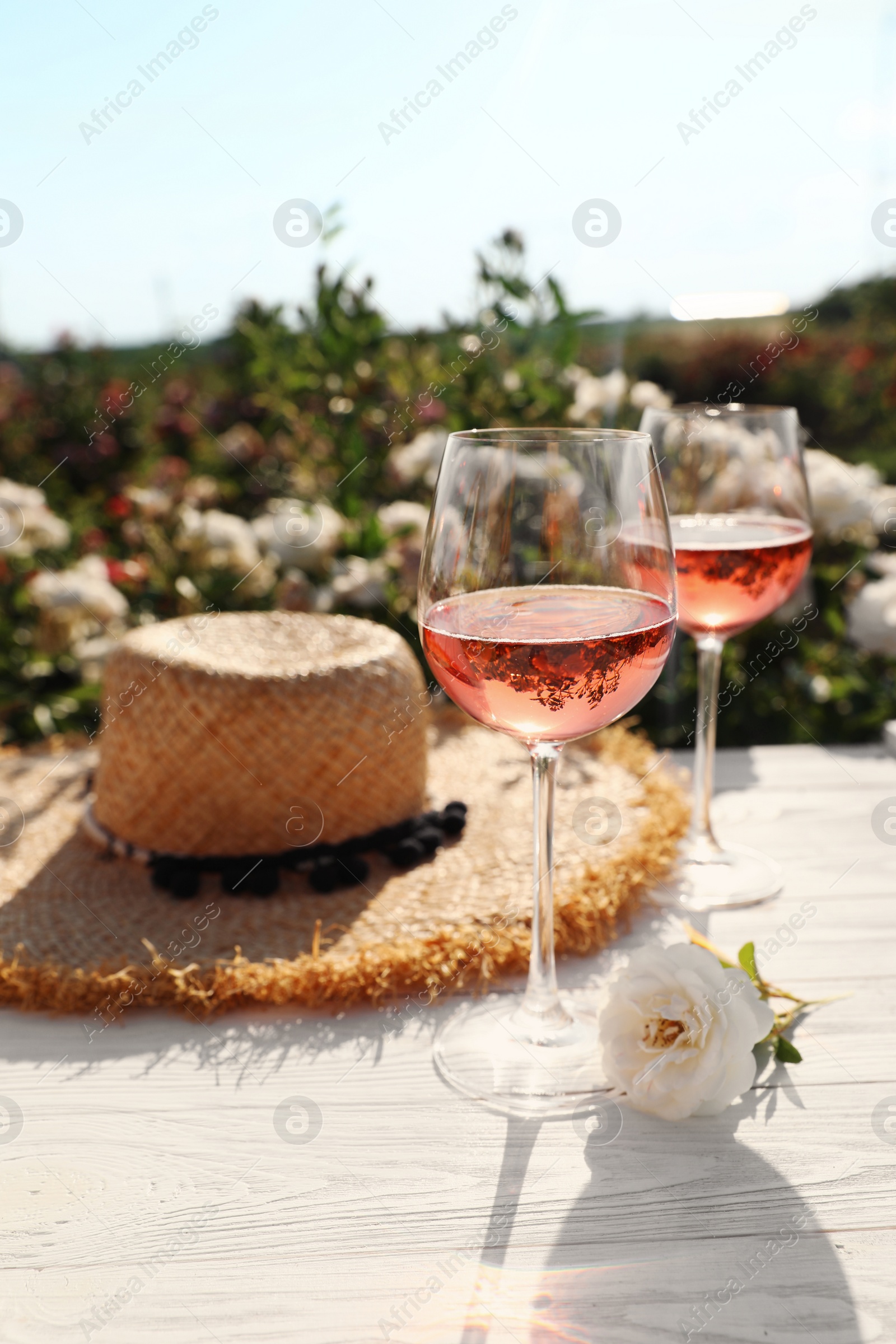 Photo of Glasses of rose wine, straw hat and beautiful flower on white wooden table outdoors