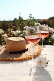 Glasses of rose wine, straw hat and beautiful flower on white wooden table outdoors