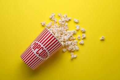 Photo of Overturned paper cup with delicious popcorn on yellow background, flat lay