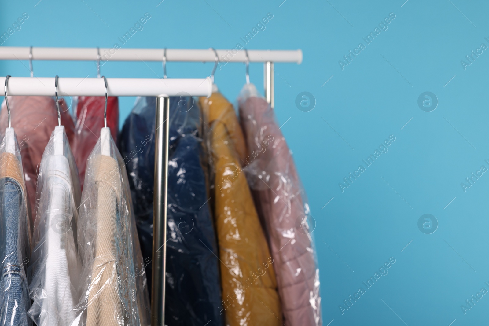 Photo of Dry-cleaning service. Many different clothes in plastic bags hanging on rack against light blue background, space for text