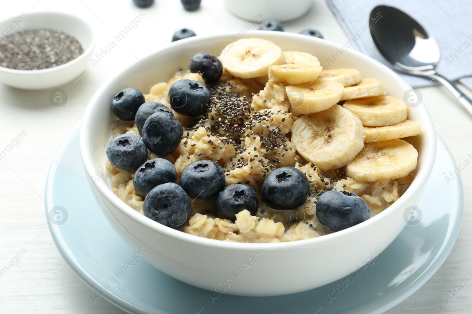 Photo of Tasty oatmeal with banana, blueberries and chia seeds served in bowl on white wooden table, closeup