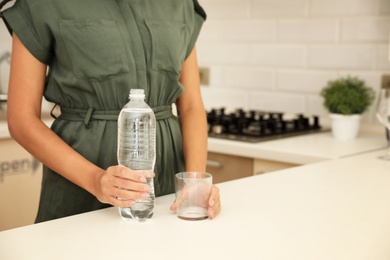 Photo of Woman with bottle and glass of water in kitchen, closeup. Space for text