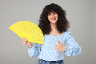 Photo of Happy woman holding hand fan and showing thumb up on light grey background