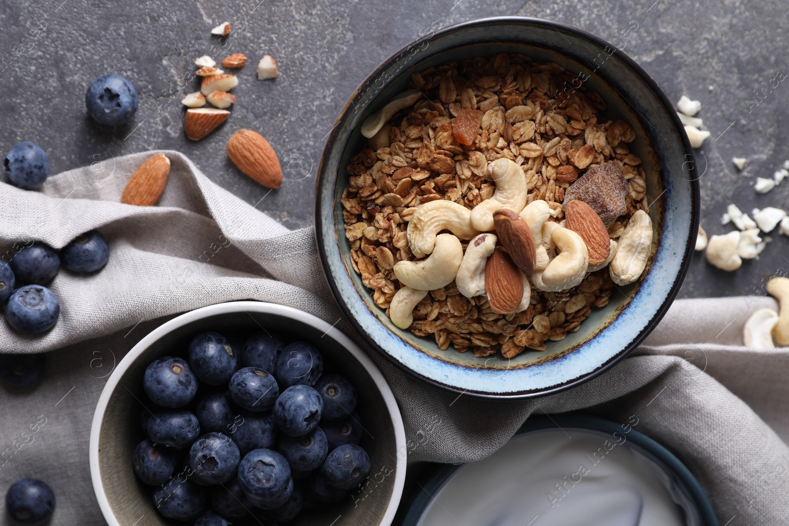 Photo of Tasty granola with nuts in bowl and blueberries on gray textured table, flat lay