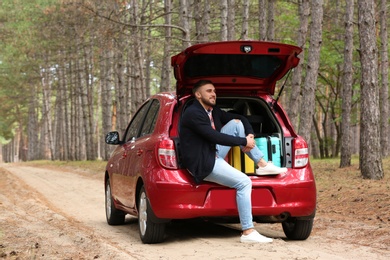 Young man sitting in car trunk loaded with suitcases on forest road
