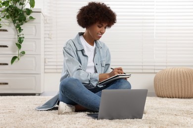 Beautiful young woman using laptop and writing in notebook in room