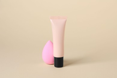 Tube of skin foundation and sponge on beige background. Makeup product