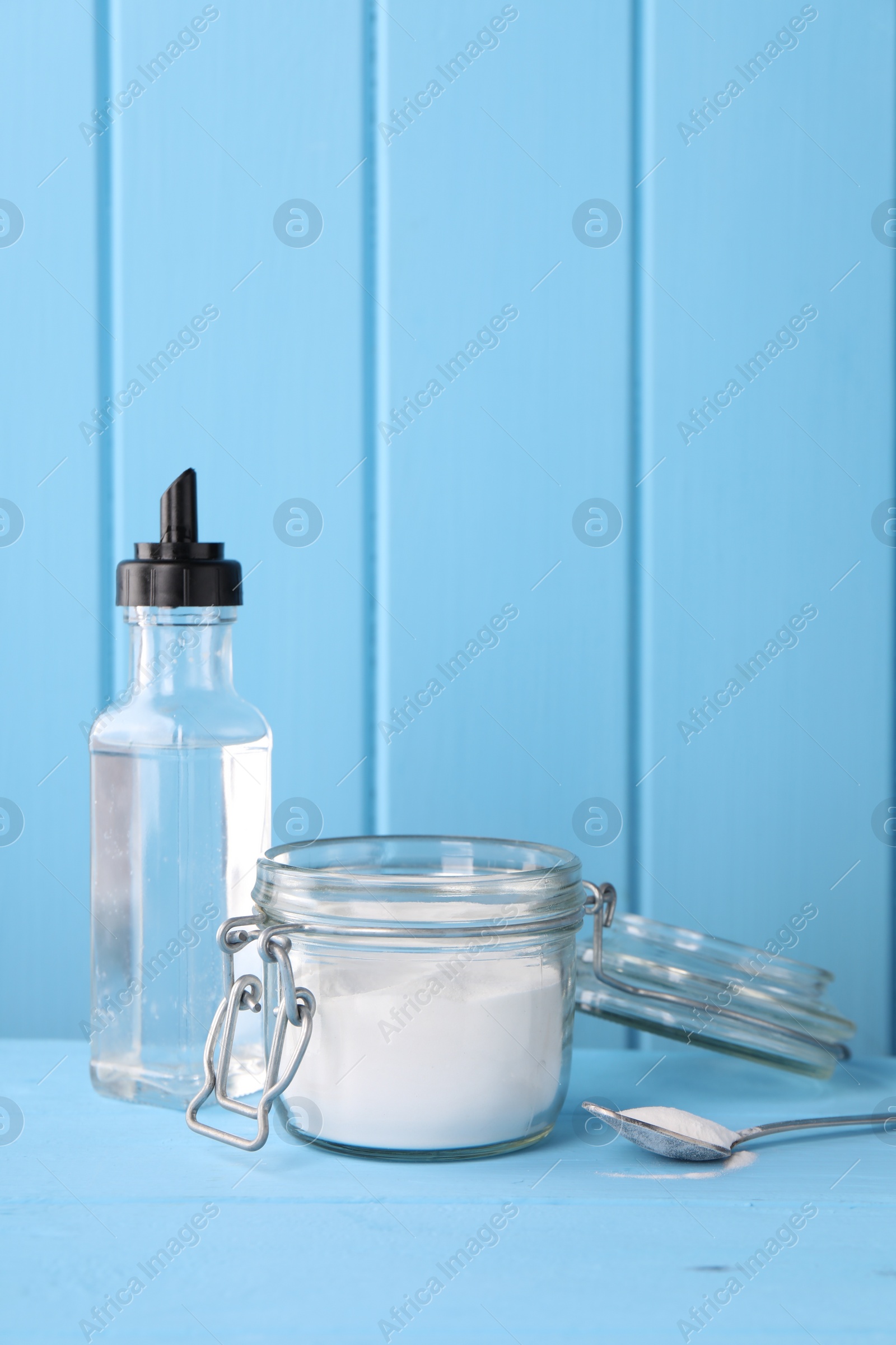 Photo of Natural cleaning products. Vinegar in bottle, baking soda and spoon on light blue wooden table