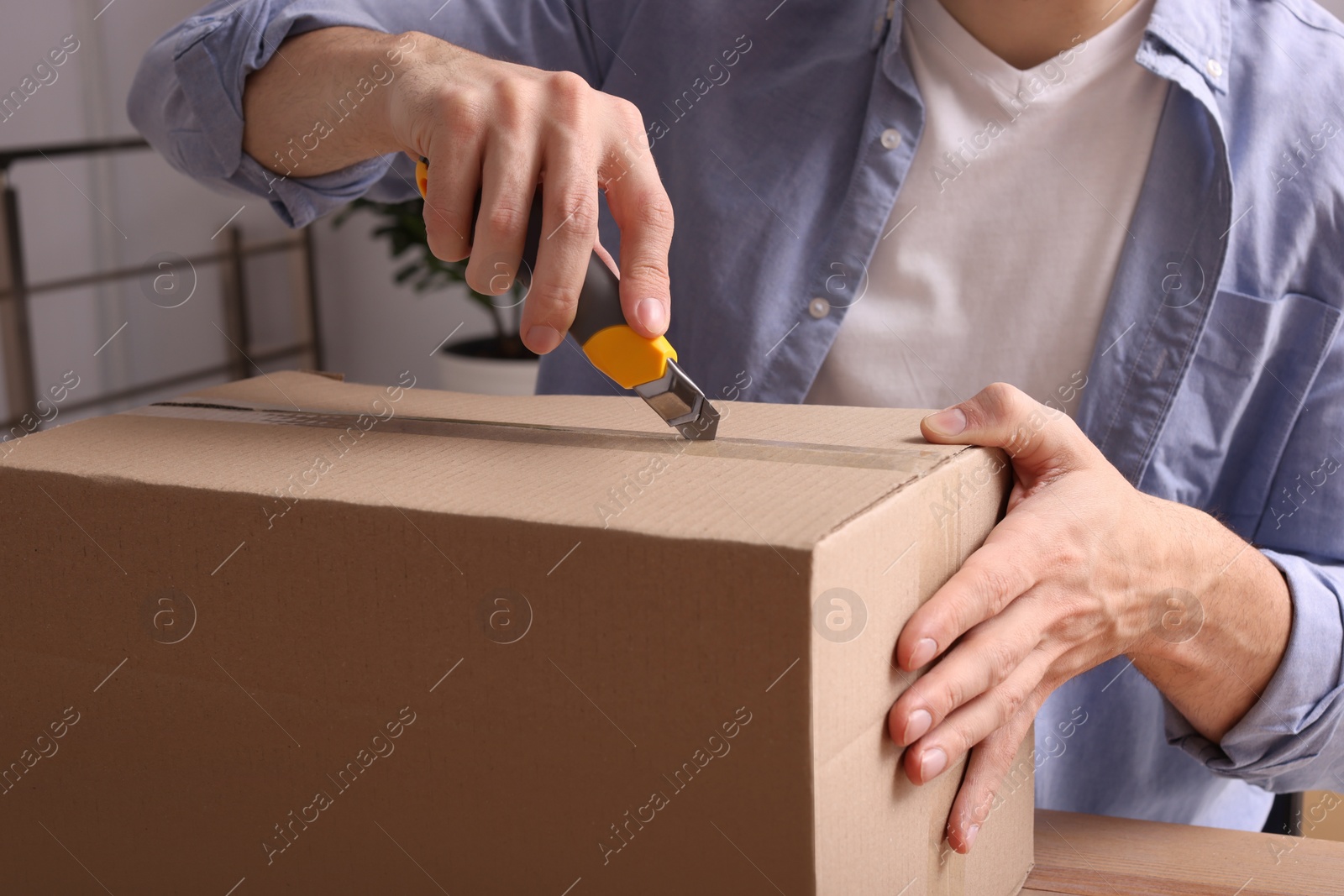 Photo of Man using utility knife to open parcel at wooden table indoors, closeup