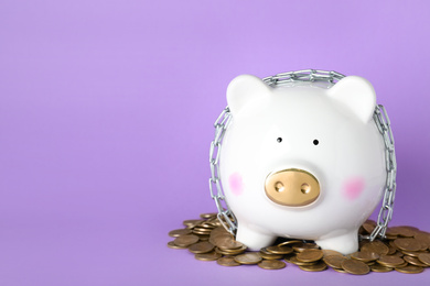 Photo of Piggy bank  with steel chain and coins and lilac background, space for text. Money safety concept