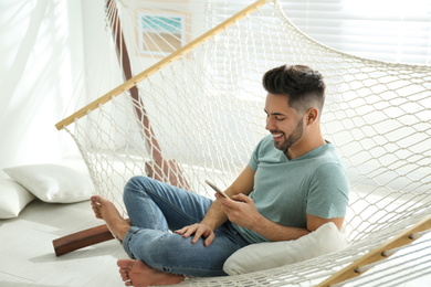 Photo of Young man using smartphone in hammock at home