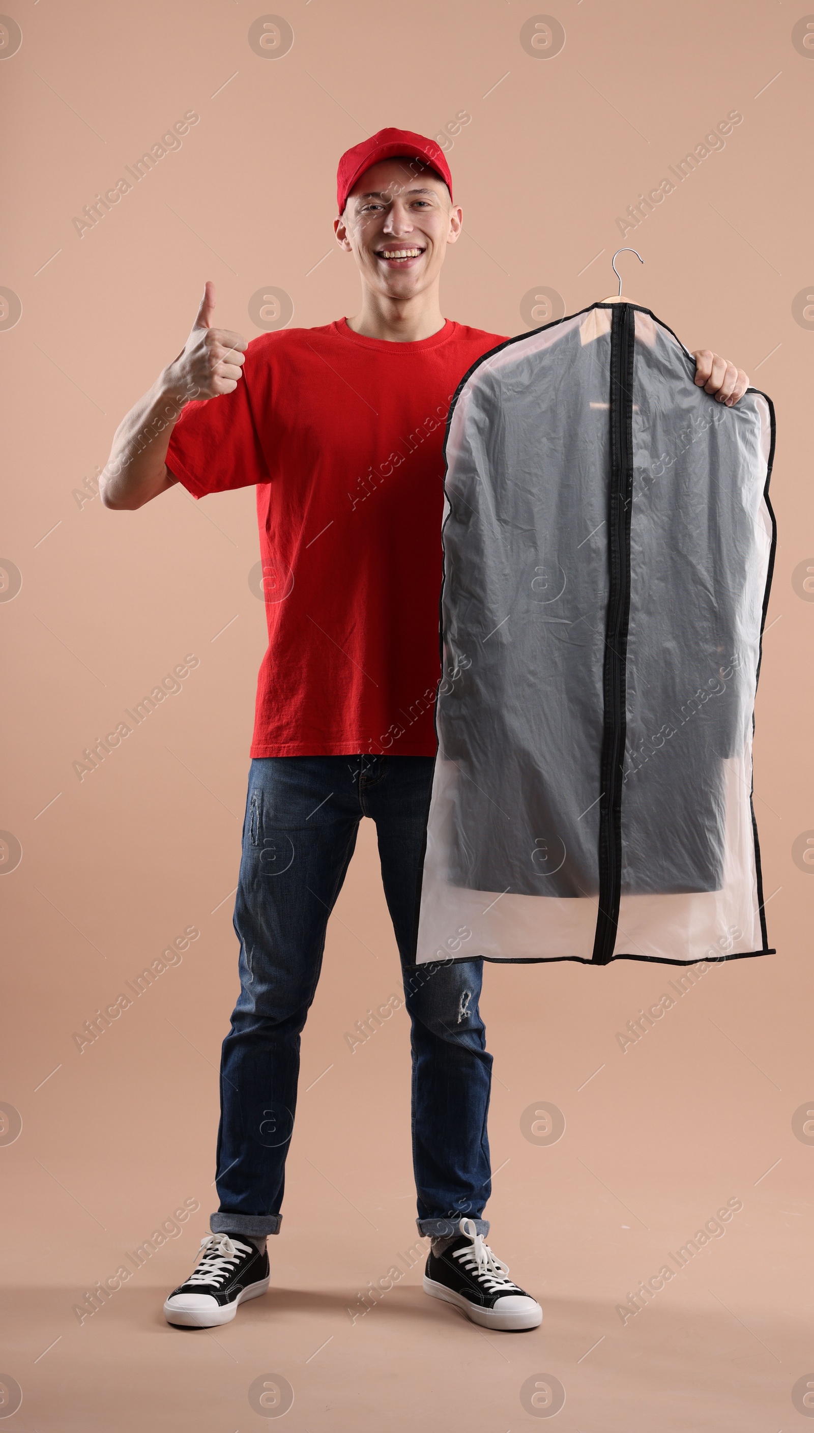 Photo of Dry-cleaning delivery. Happy courier holding garment cover with clothes and showing thumbs up on beige background