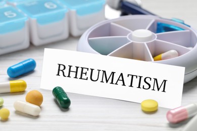 Photo of Card with word Rheumatism and pills on white wooden table, closeup