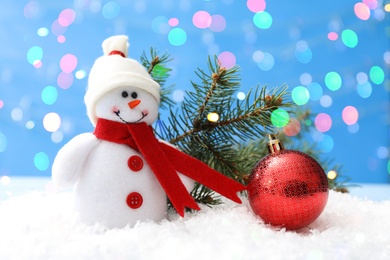 Image of Cute snowman, Christmas ball and fir tree branch on snow against light blue background, bokeh effect