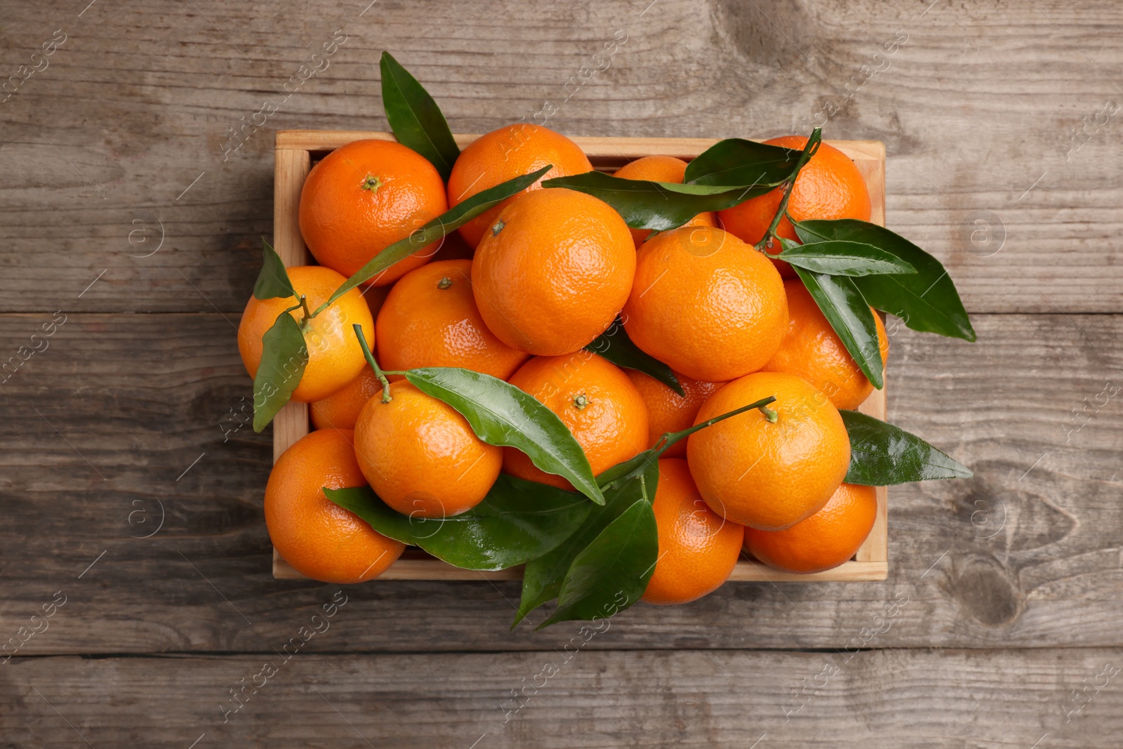 Photo of Delicious tangerines with leaves in crate on wooden table, top view