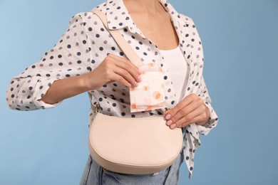 Photo of Young woman putting menstrual pad into bag on light blue background, closeup