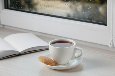 Photo of Cup of tea, saucer with cookie and open book on wooden window sill