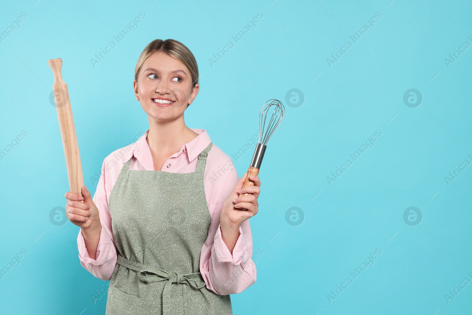 Photo of Beautiful young woman in clean apron with kitchen tools on light blue background. Space for text