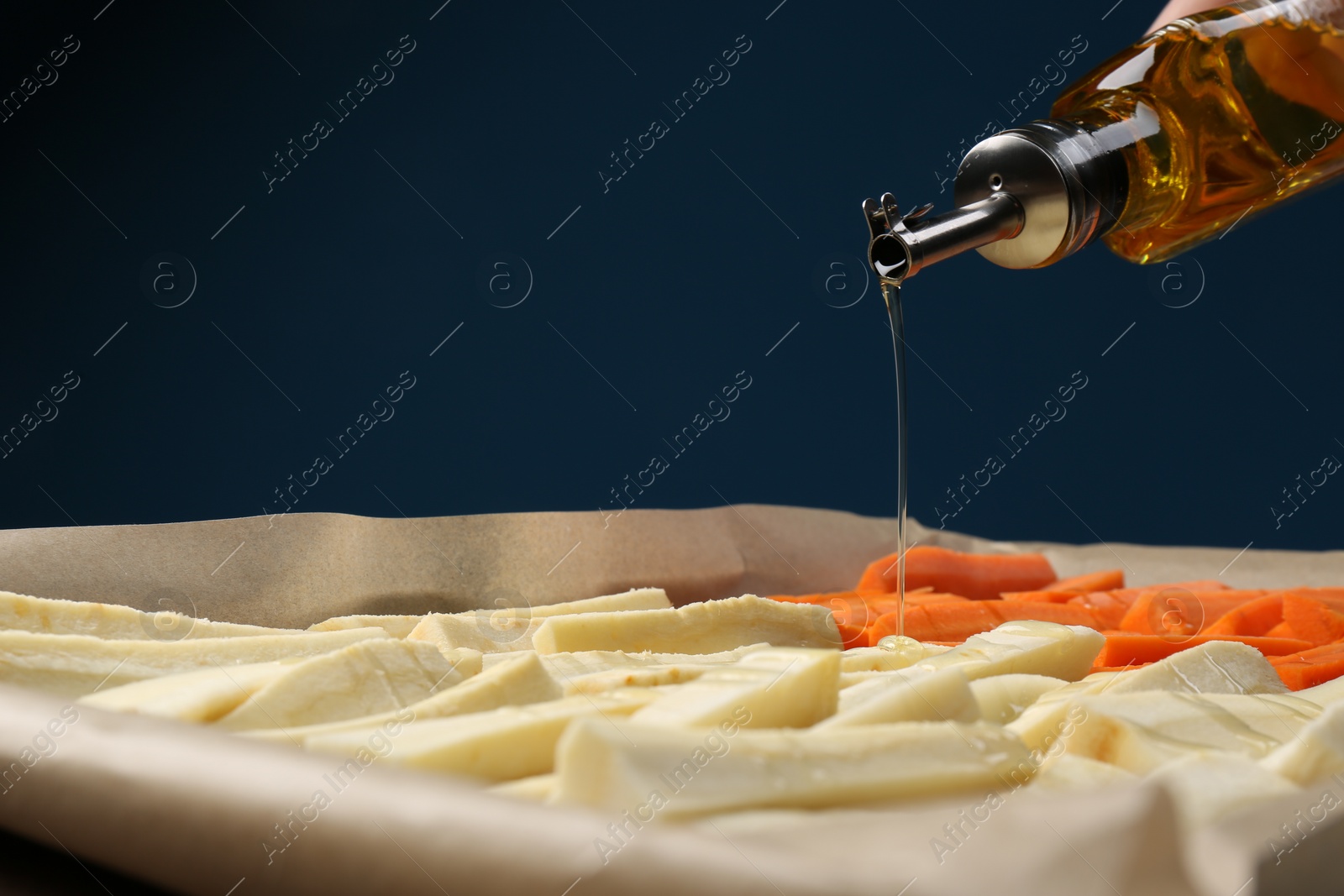 Photo of Pouring oil onto baking tray with parsnips and carrots against blue background, closeup