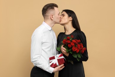 Lovely couple with gift box and bouquet of red roses kissing on beige background. Valentine's day celebration
