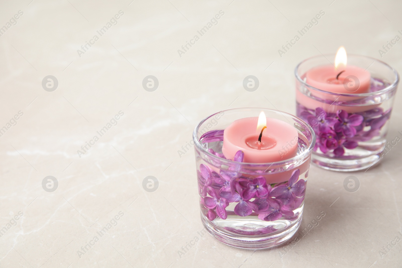 Photo of Glass holders with water, lilac flowers and burning candles on light table, space for text