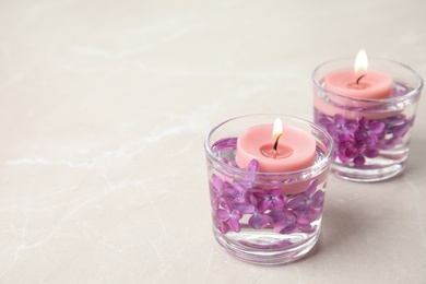 Photo of Glass holders with water, lilac flowers and burning candles on light table, space for text