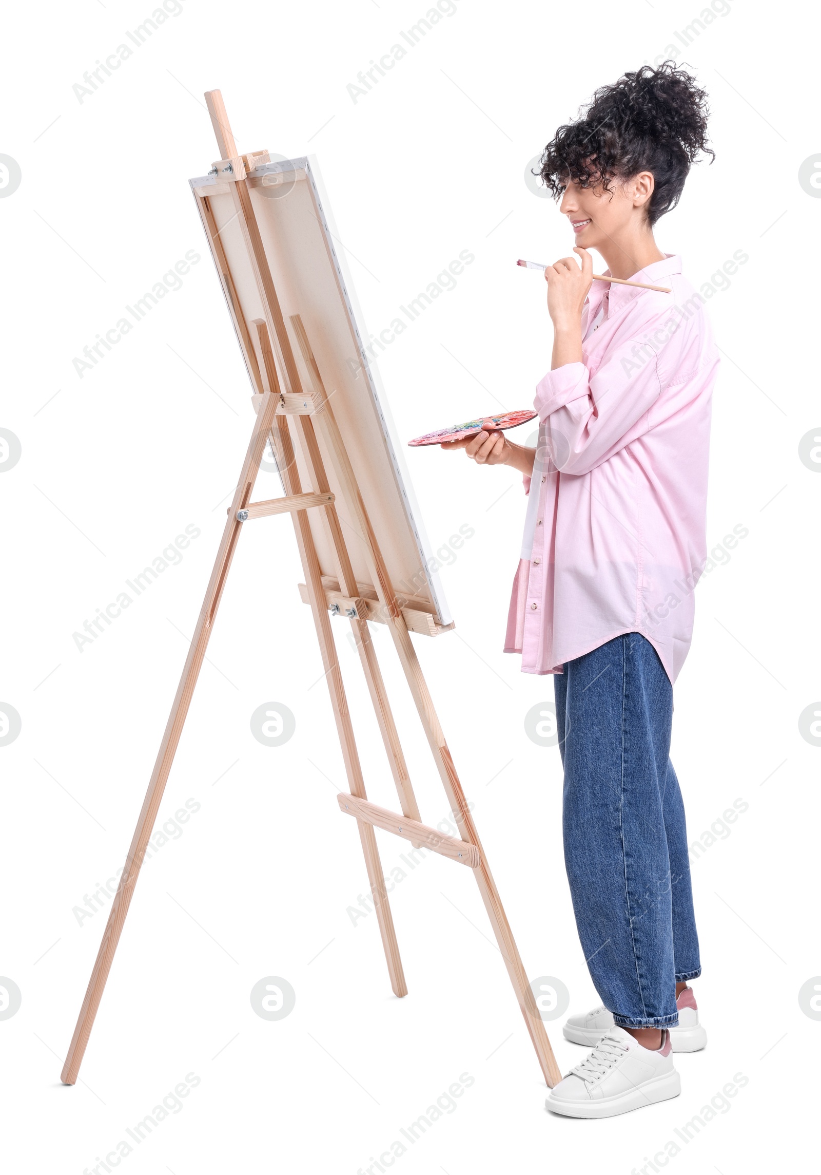 Photo of Young woman holding brush and artist`s palette near easel with canvas against white background