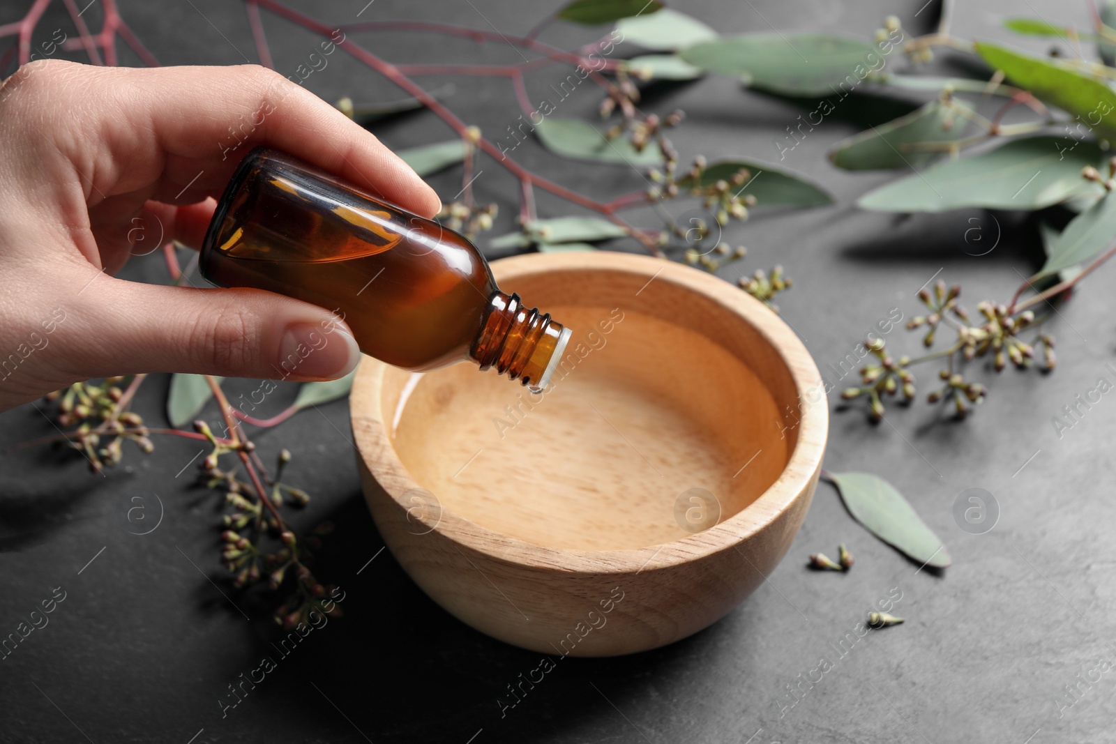 Photo of Woman dripping eucalyptus essential oil from bottle into bowl at grey table, closeup