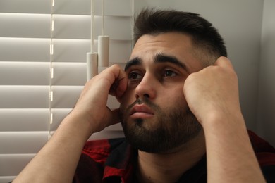 Sad young man near closed blinds at home