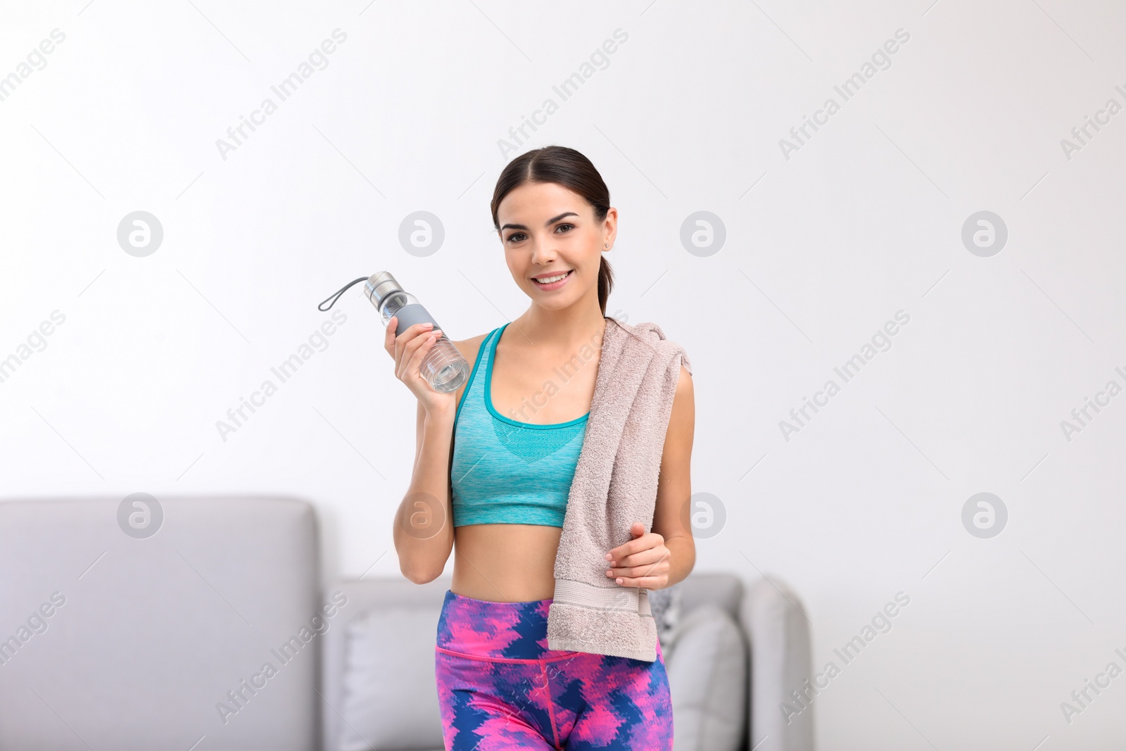 Photo of Beautiful young woman in sportswear with towel and bottle of water indoors