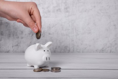 Woman putting coin into ceramic piggy bank at white wooden table, closeup with space for text. Financial savings