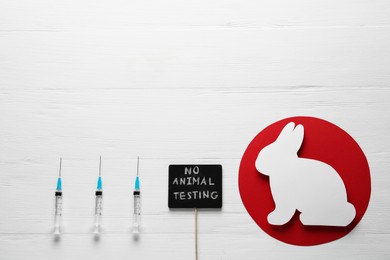 Signboard with text No Animal Testing, figure of rabbit and syringes on white wooden table, flat lay