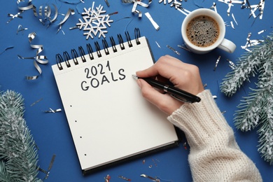 Photo of Woman writing in notebook on blue background with Christmas decor, top view. New year aims