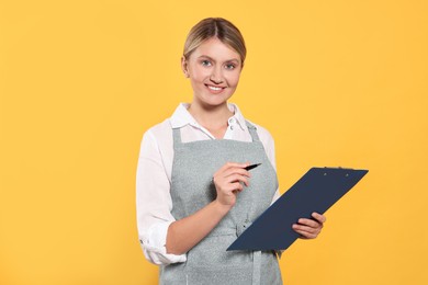 Photo of Beautiful young woman in clean apron with clipboard on orange background